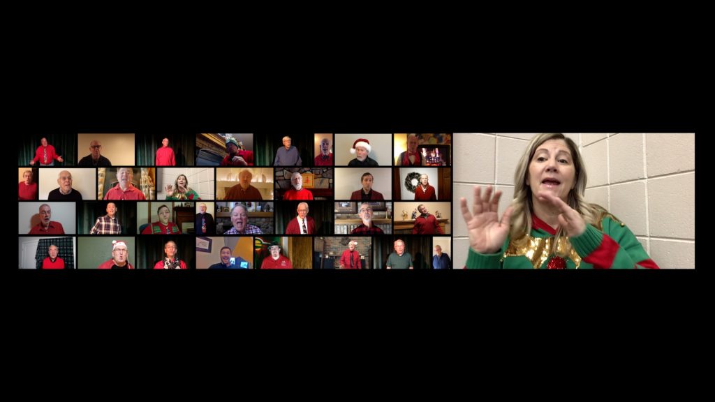 A multi-screen grid where each rectangle features a different singer in a 31-man chorus. Everyone is wearing festive holiday colors. Some of the frames are vertical, and some are horizontal. The background is black. There is a large frame to the right of the grid which features the music director, Jayme Casperson, who is using her hands to direct with passion while singing along.