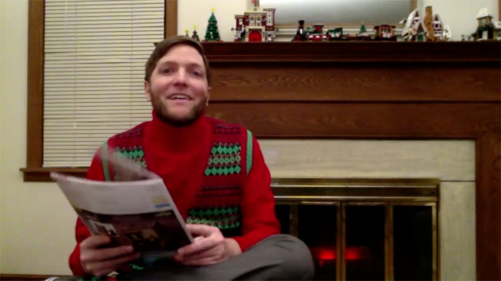 A picture of Blake Wanger, a white man with light brown hair and a beard, who is smiling at the camera. He is wearing a red Christmas sweater with green patterns, and holding a magazine. Behind him is a fireplace with Christmas-themed LEGOs on the mantle.